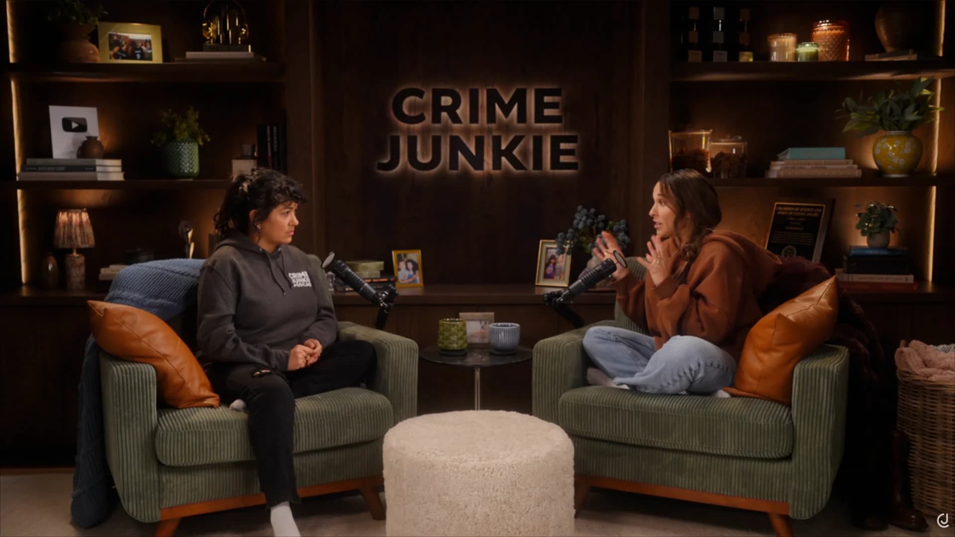 Two hosts recording in armchairs in the Crime Junkie studio, with microphones and a wood shelving backdrop.